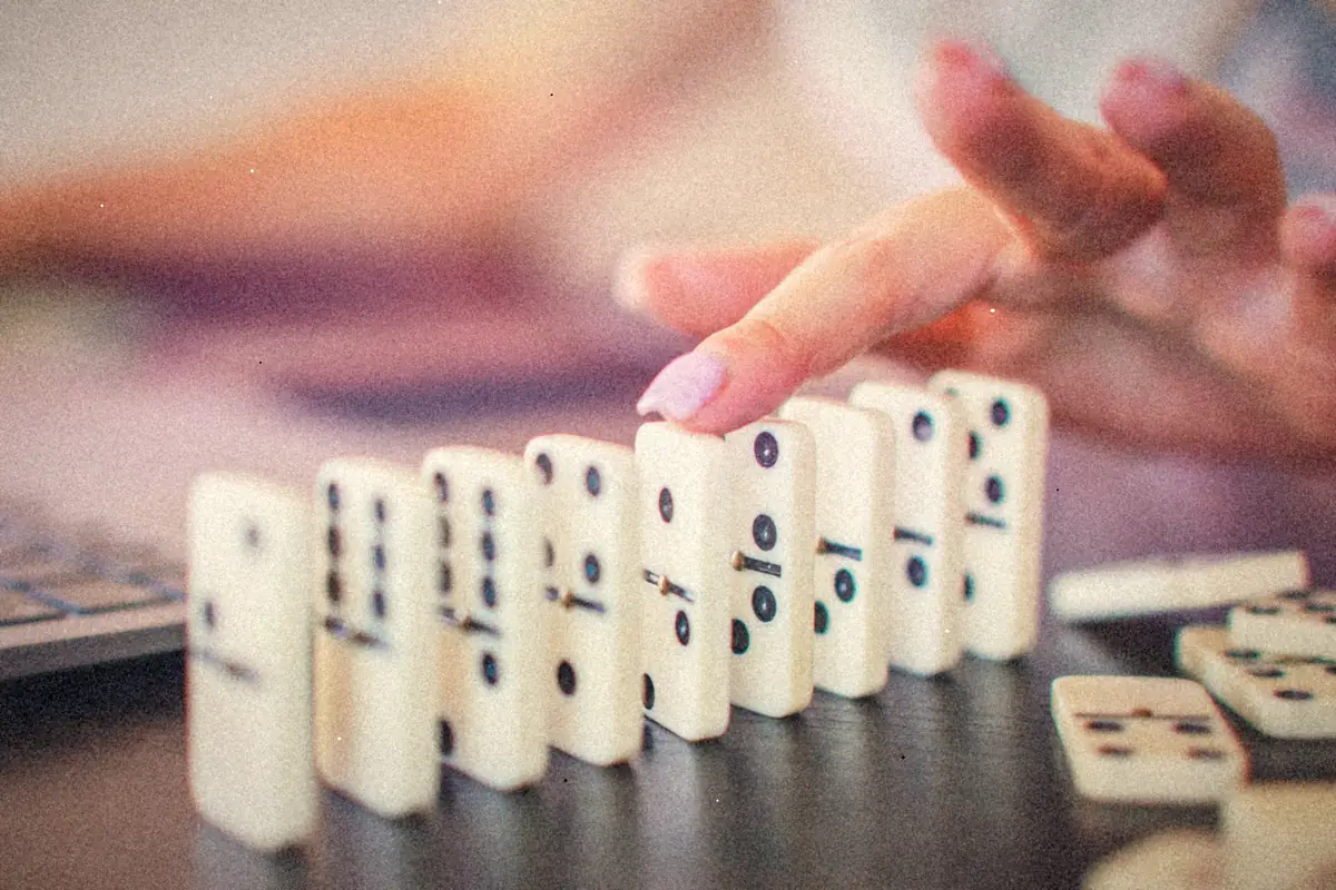 A finger tips the first domino in a line of white dominoes on a table.