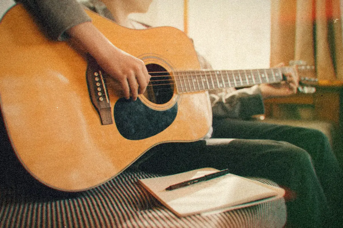 Someone playing an acoustic guitar with a notebook and pen close by.