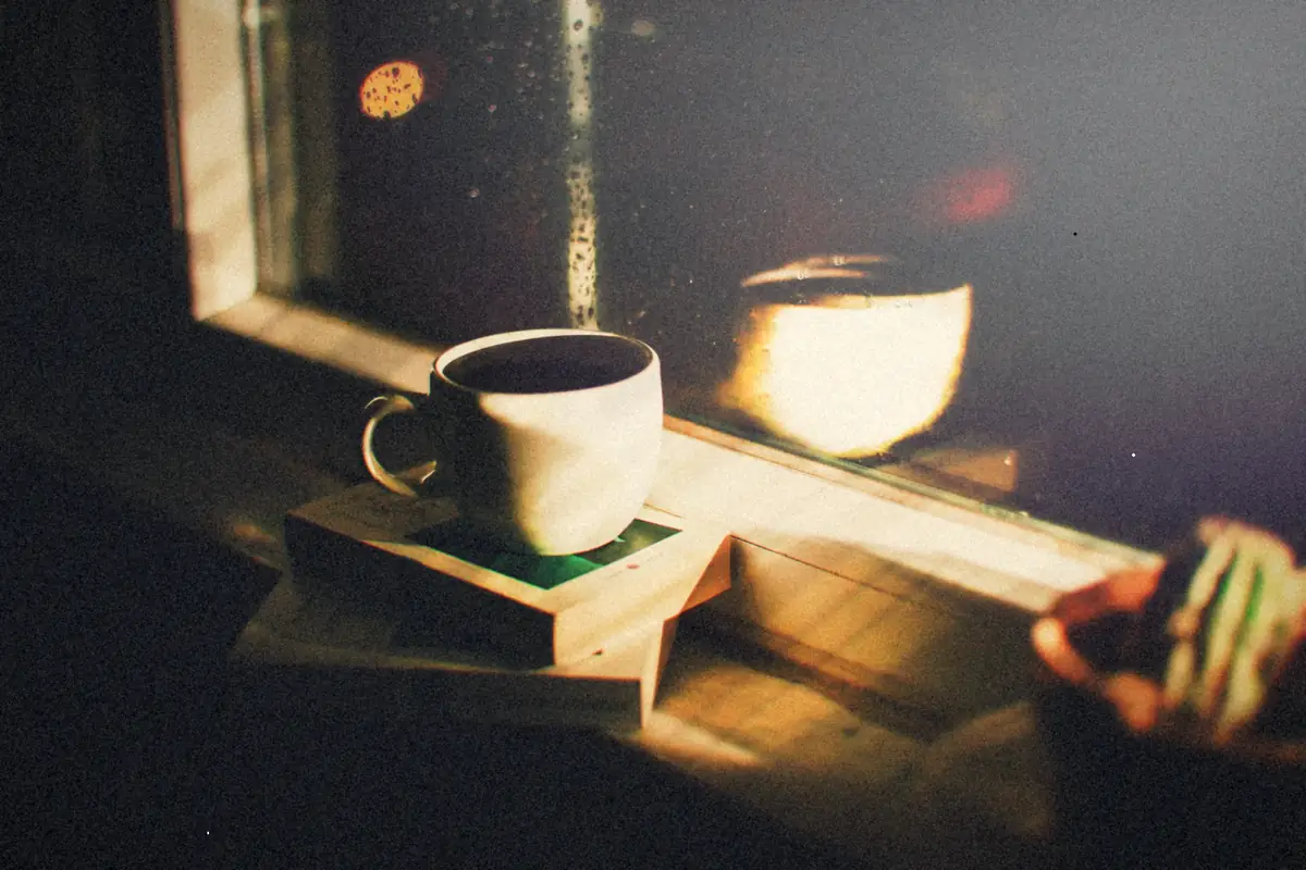 A cup of coffee on a stack of books by a rain-streaked window at night.