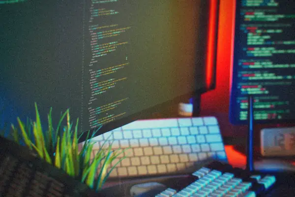 A developer's desk with code on two monitors, a mechanical keyboard, a small clock and a desk plant.